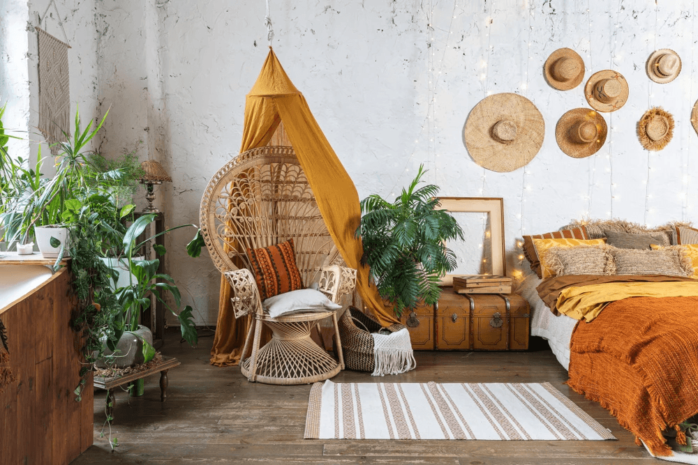 Boho-Raum mit Rattansessel, Hängepflanzen, Strohhüten an der Wand, Holzmöbeln und senfgelber Bettdecke